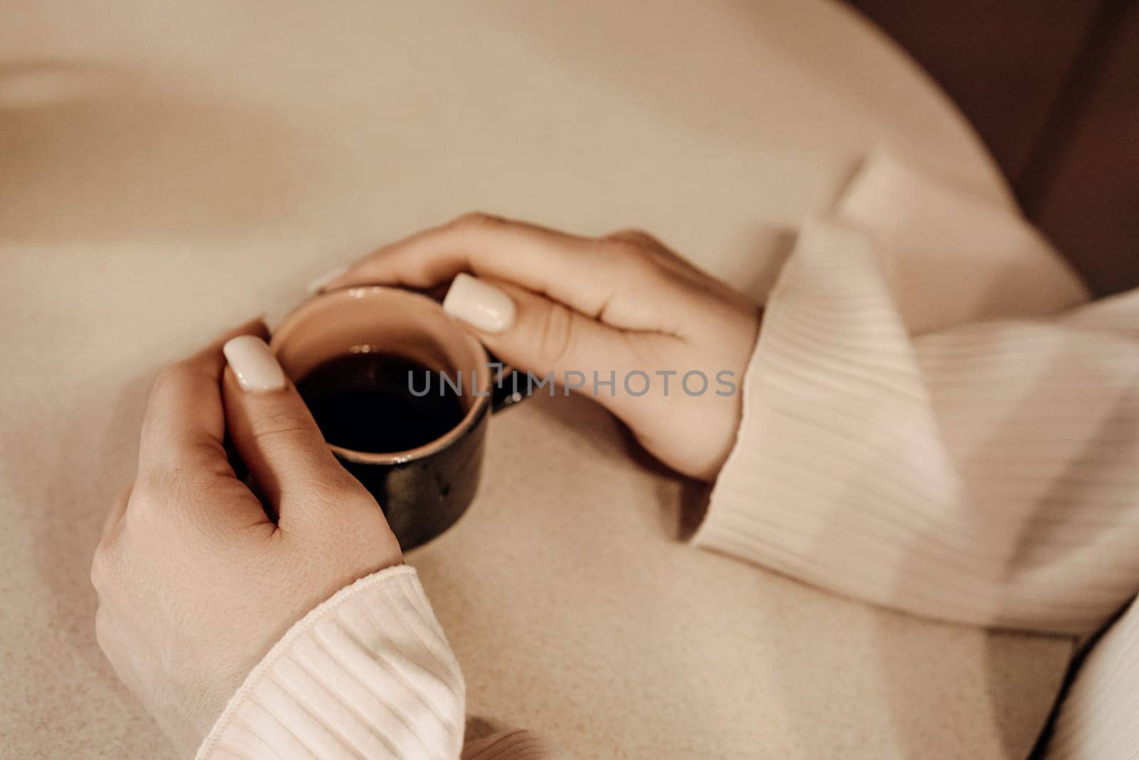 Two hands gently cradle a small, dark espresso cup. Beige sweater sleeves and a neutral background. Warm, intimate setting.