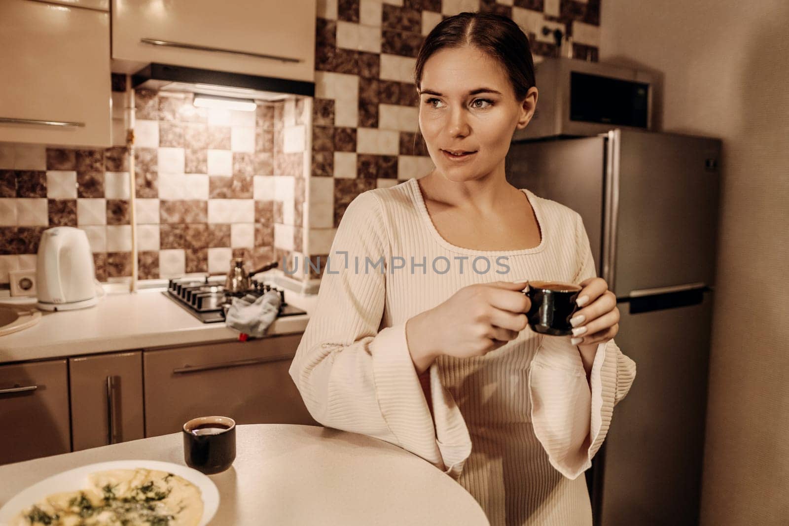 Woman in white robe stands in kitchen, holding small dark mug. A plate of food and a coffee pot are on the counter. She looks thoughtfully away.