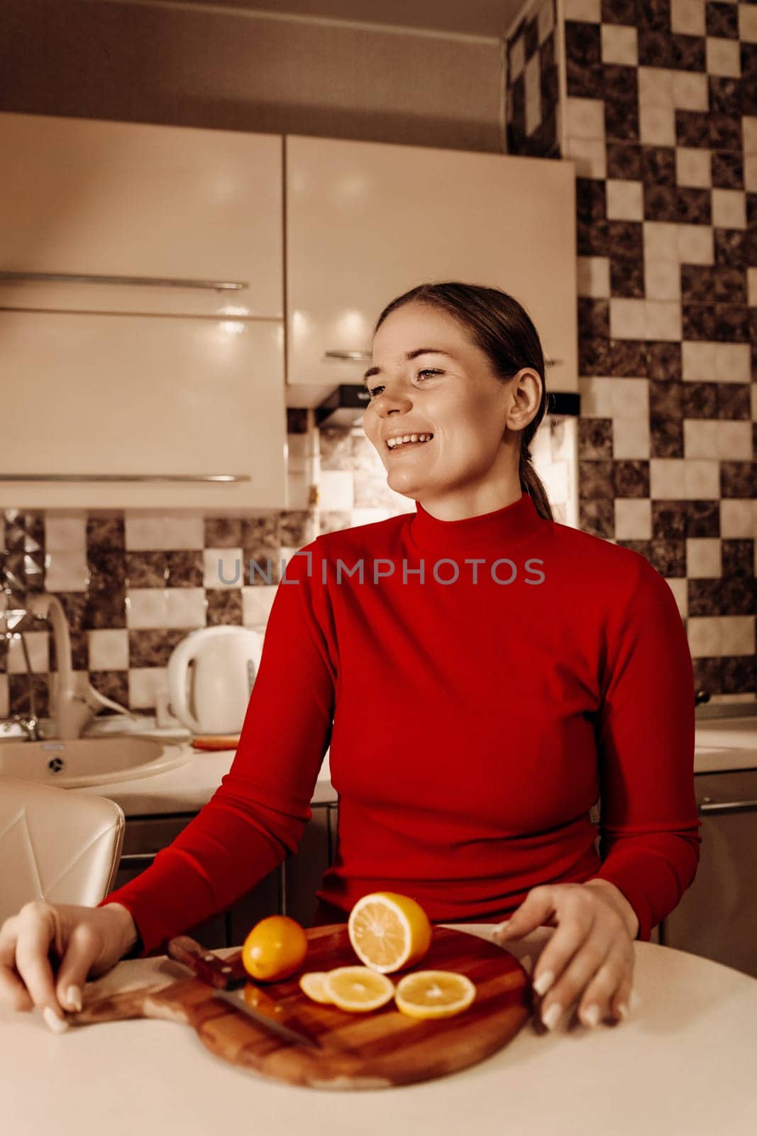 Smiling woman in a red turtleneck sits at a kitchen counter, a wooden board holding sliced oranges and a lemon before her.