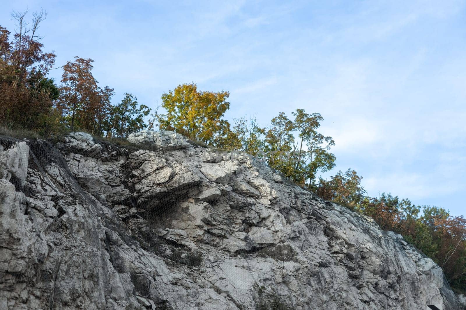 Rocky cliff with patches of green and autumn trees under a partly cloudy blue sky depicting natural erosion and geology.