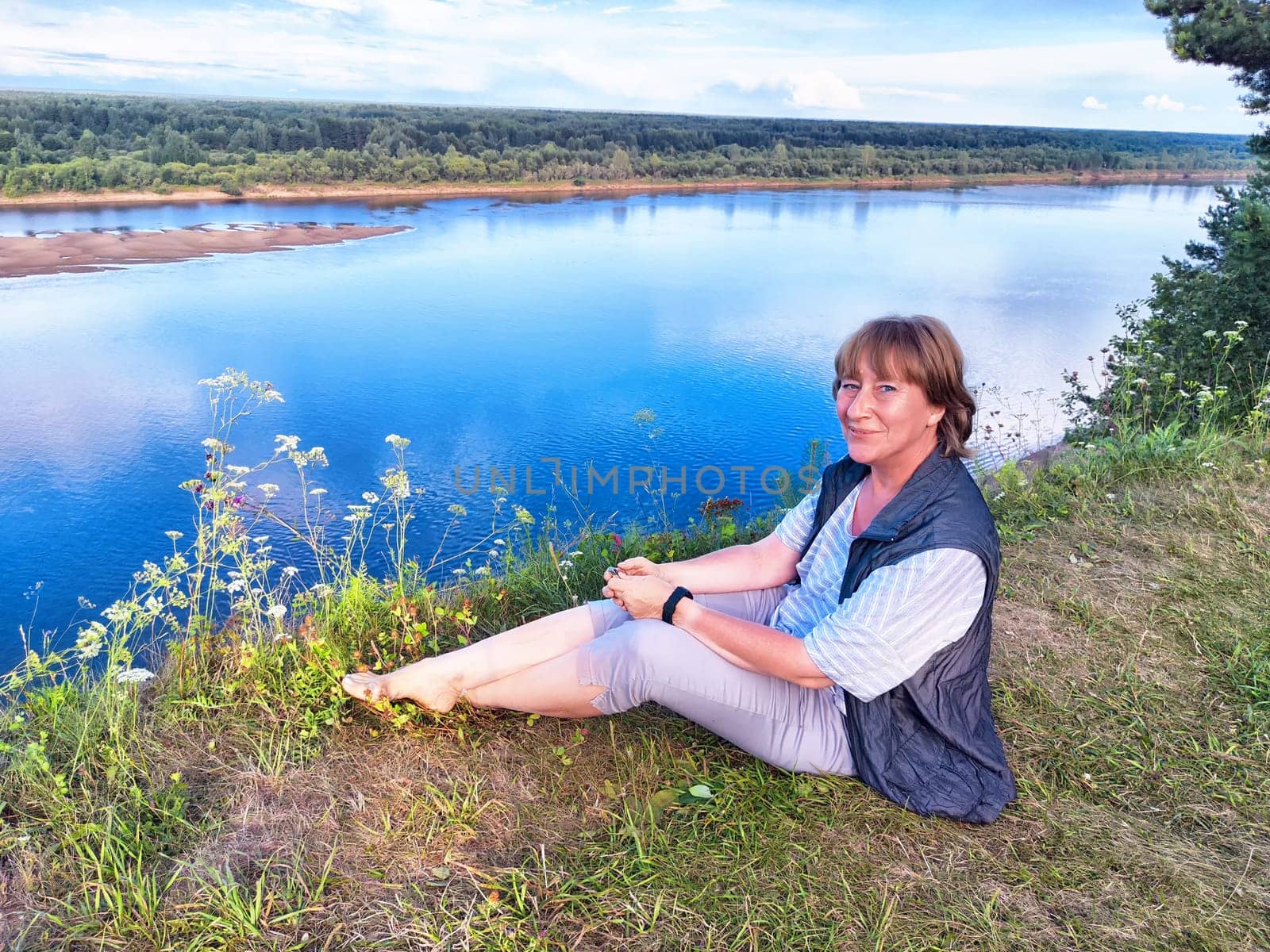 A middle-aged female traveler blogger captures a selfie by a serene river, surrounded by lush greenery.