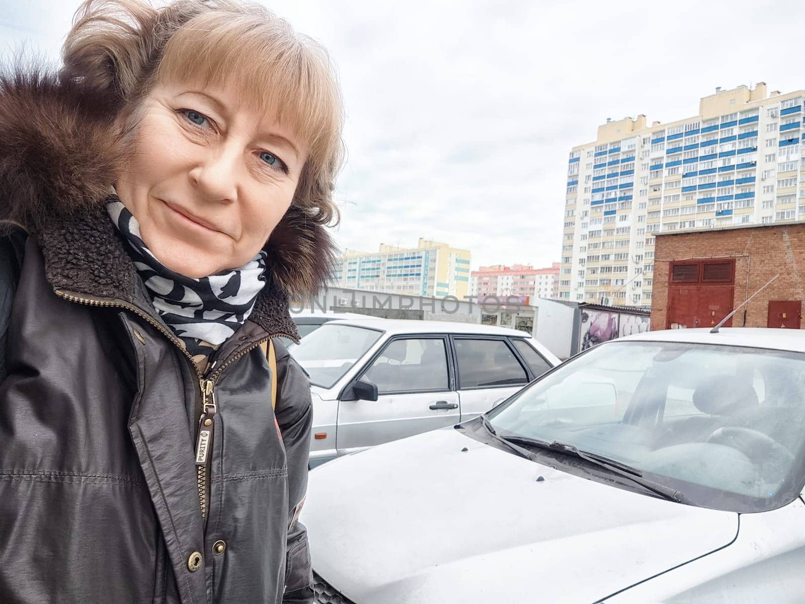 A woman stands smiling next to a parked car, showcasing urban surroundings with residential buildings and clouds.