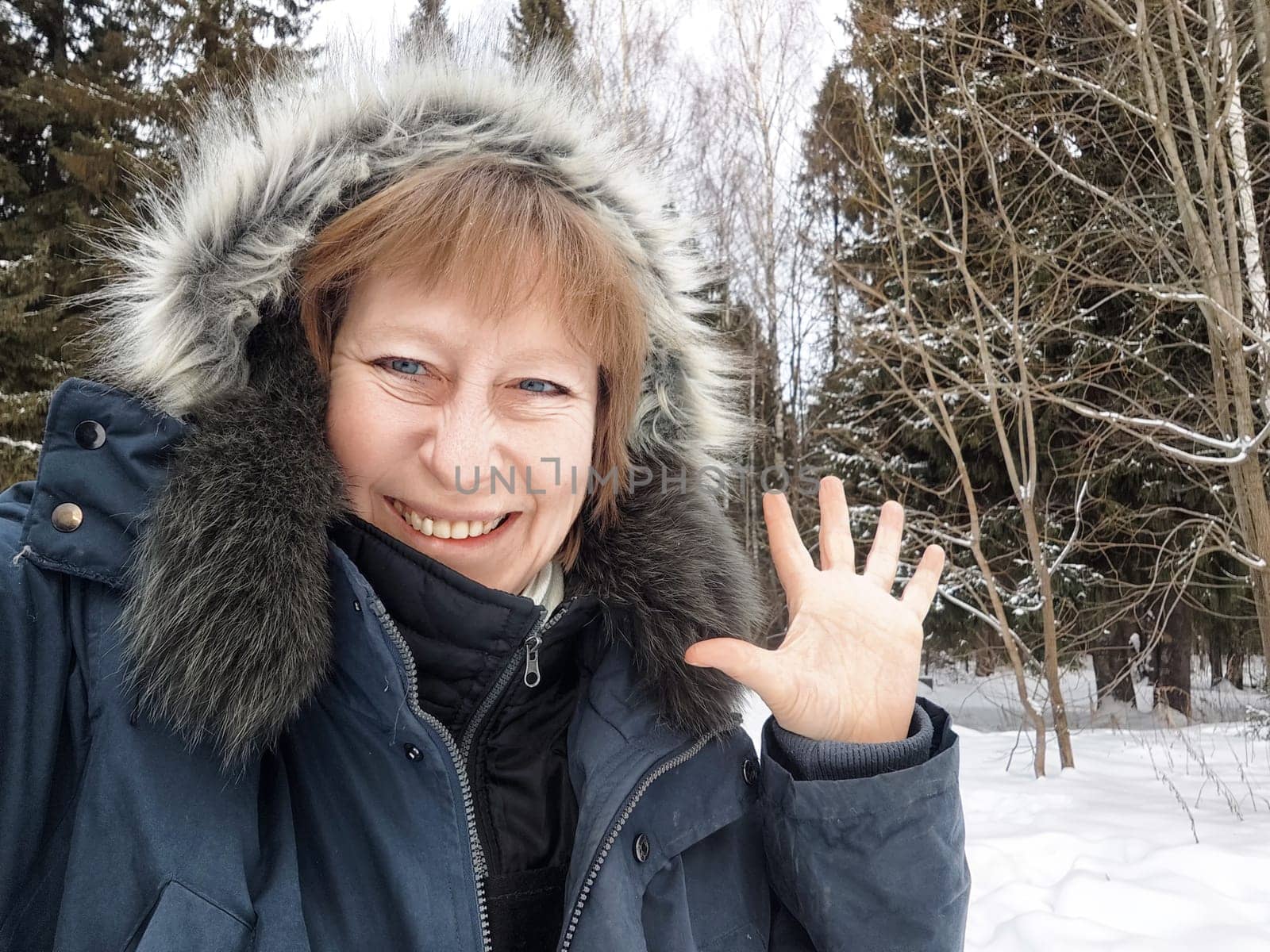 A middle-aged blonde woman dressed warmly greets the camera with a smile in a snowy forest during winter.
