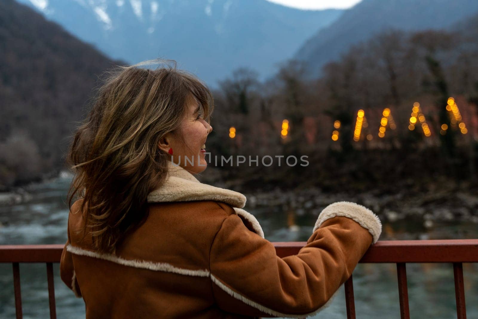 Woman in brown suede jacket gazes at illuminated structures nestled in a mountain valley, a river flowing below. Autumnal landscape.