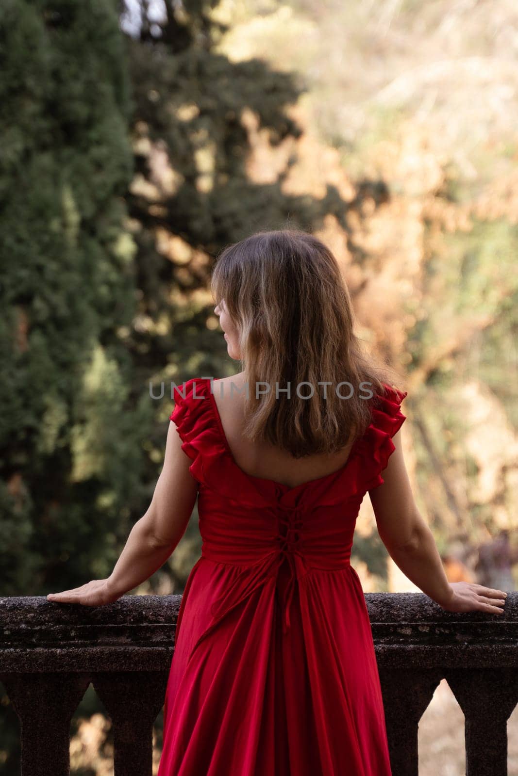 Woman in a red dress stands on a balcony overlooking a lush, autumnal landscape. Her back is to the camera.