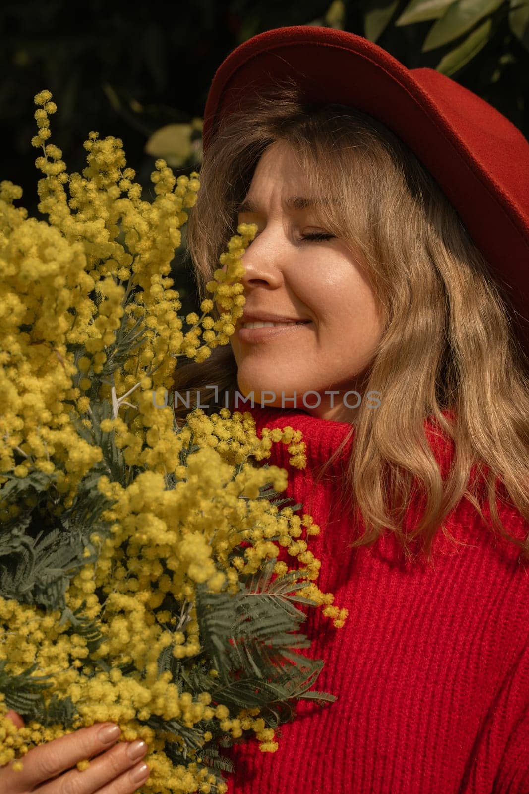 Blond woman in red hat and sweater, eyes closed, smiling, deeply inhaling the scent of a large bouquet of bright yellow mimosa flowers.