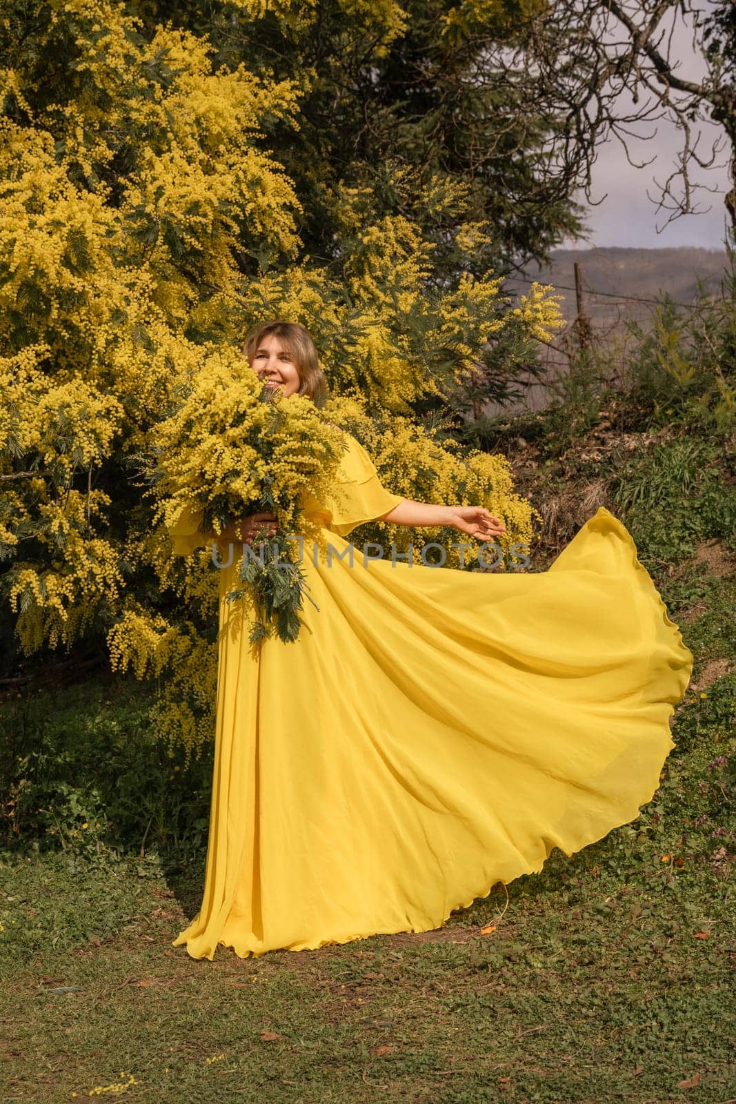 Woman in flowing yellow gown holds mimosa blossoms near a mimosa bush. Sunlight illuminates the scene. Dress billows gently.