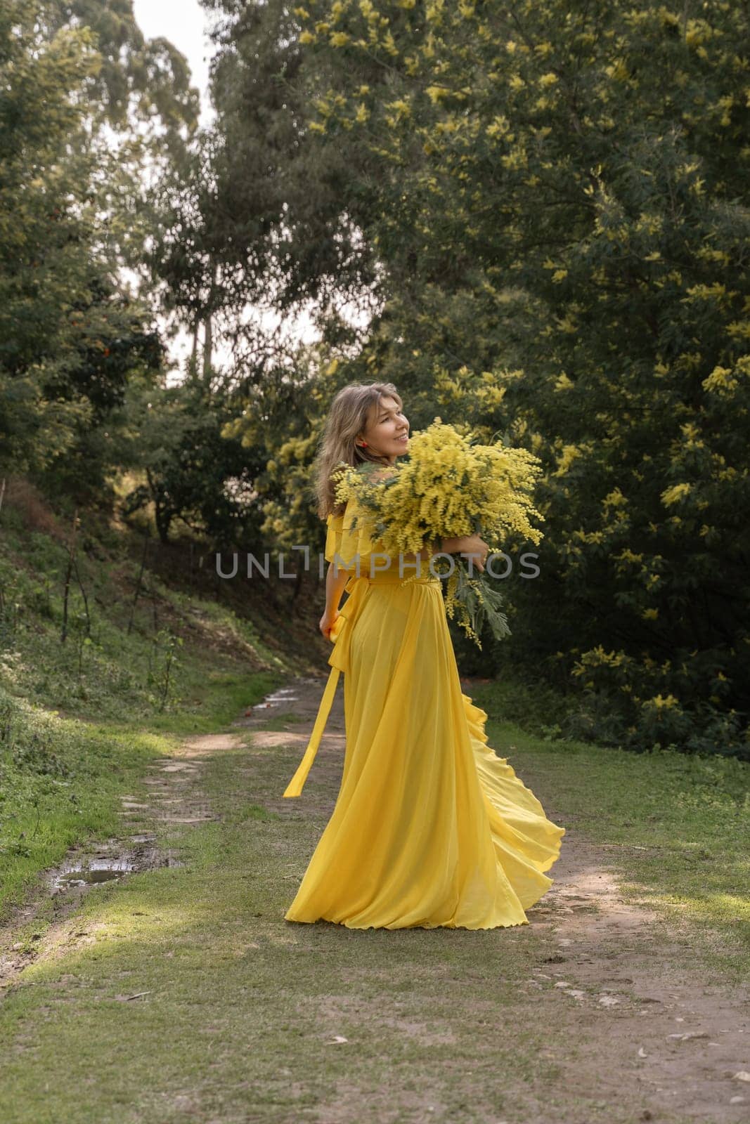 Woman in a flowing yellow maxi dress holds a large bouquet of yellow flowers while standing on a path in a wooded area.