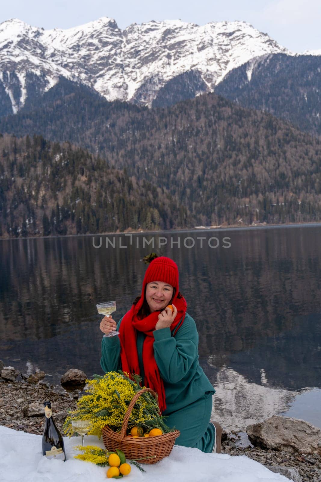 Woman enjoys a lakeside picnic amidst snowy mountains. A basket of fruit and flowers sits nearby. She holds a glass, possibly wine. Serene winter landscape.