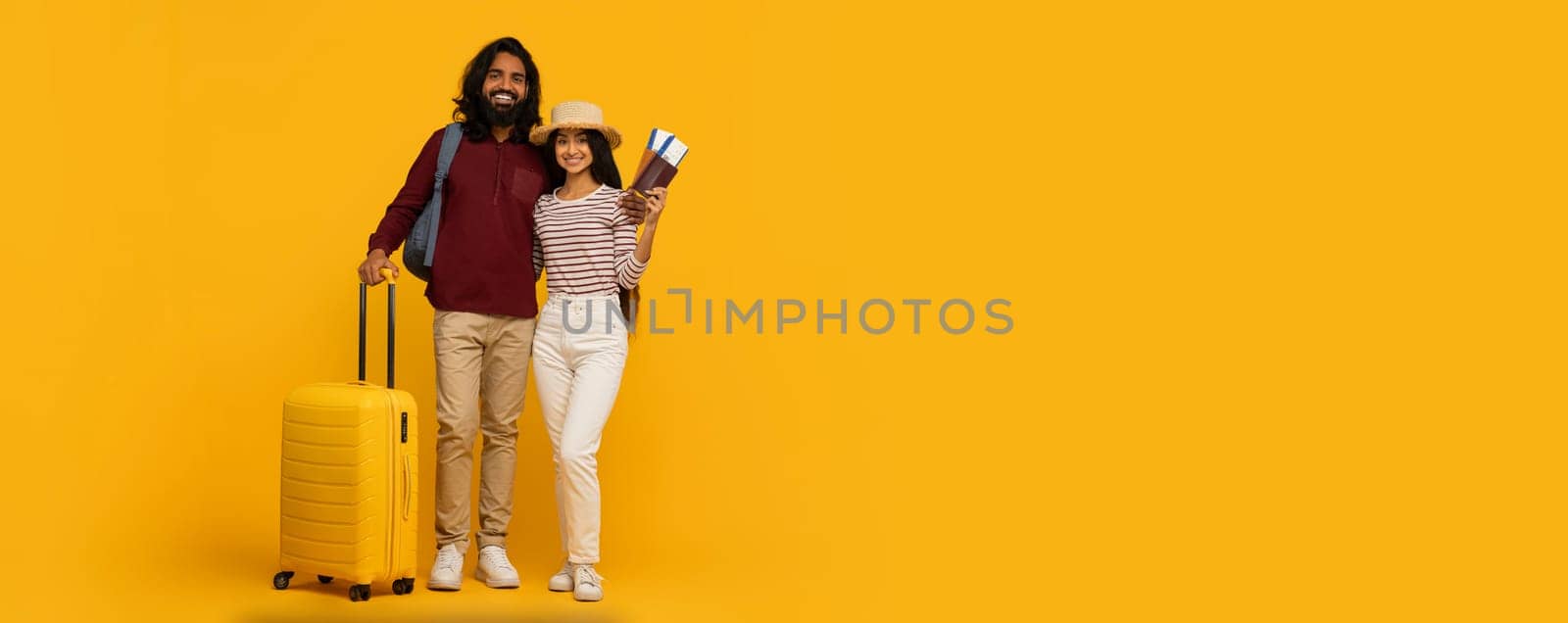 Happy traveling couple standing close with passports and a yellow suitcase on a sunny yellow background, copy space