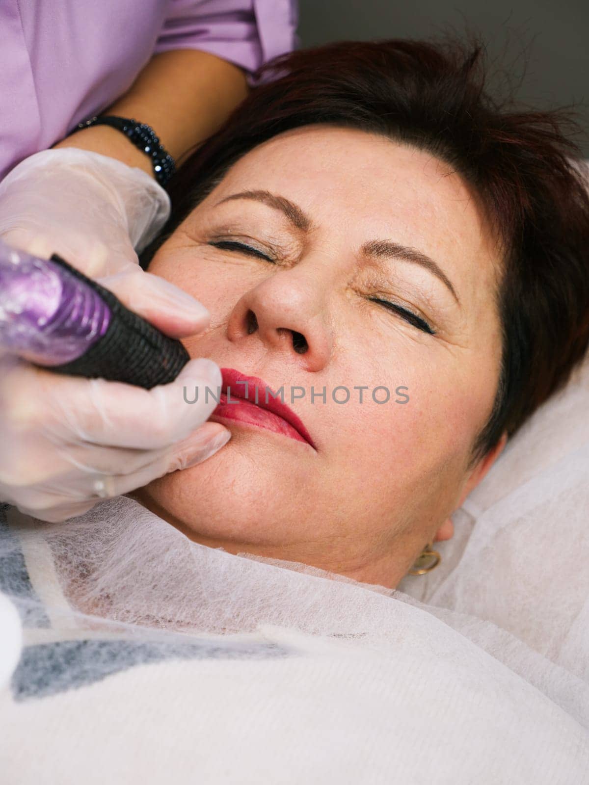 Permanent lip makeup beautiful woman over 50 years old in a beauty salon. A close-up beautician does a tattoo on his face. Vertical
