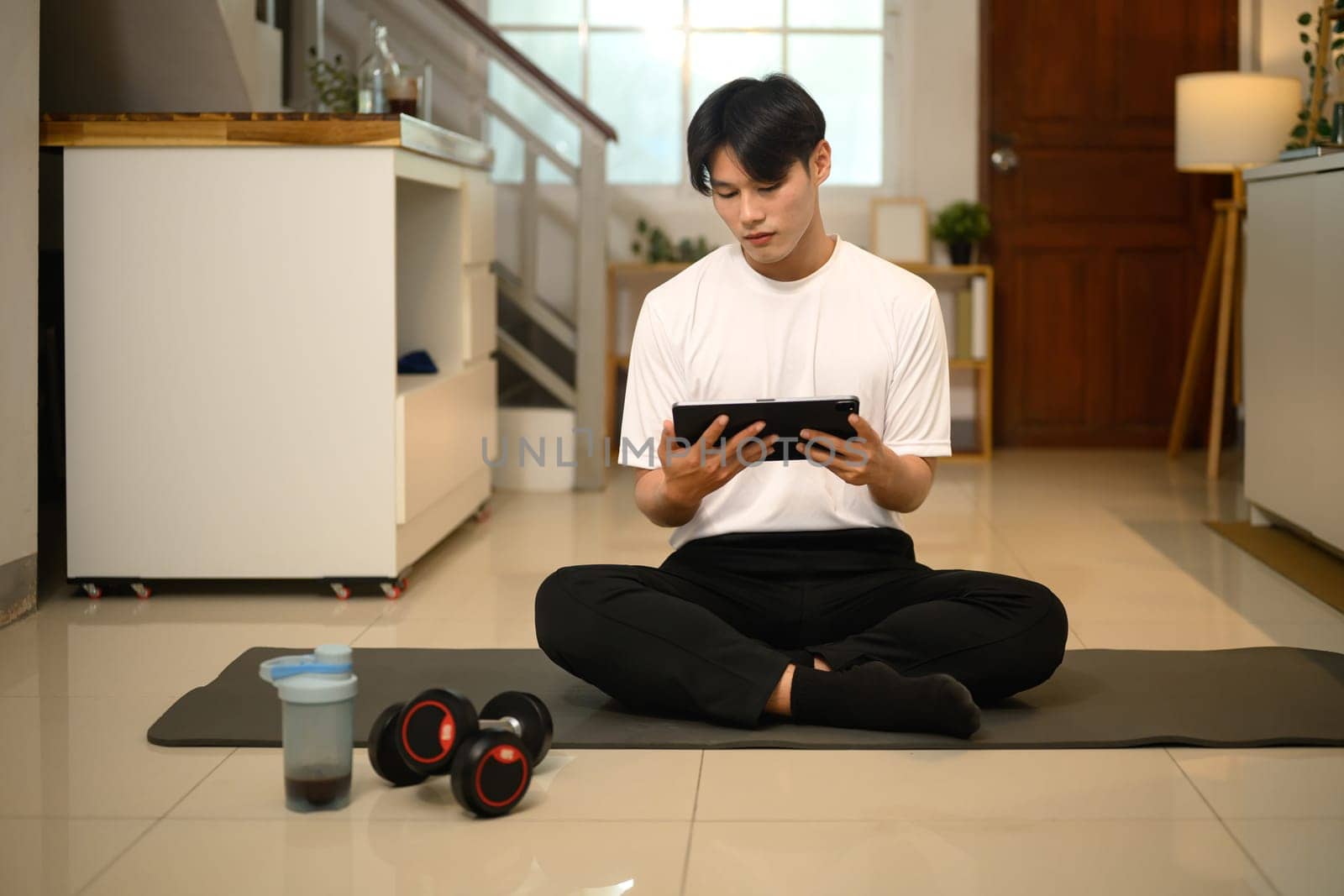 Young asian man sitting on yoga mat and using digital tablet preparing for home workout.