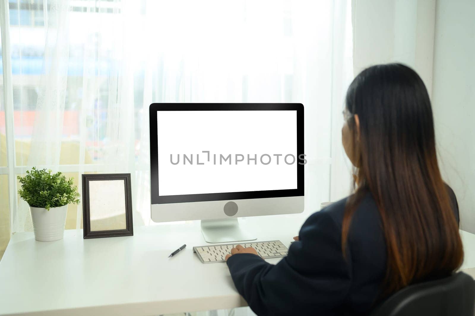 Back view of businesswoman sitting at desk and focused on desktop computer with a blank white screen.