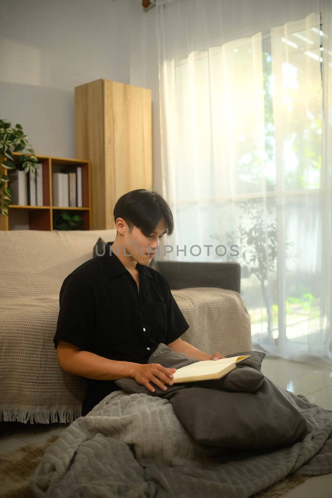 Calm asian man sitting on floor in bright living room and reading book.