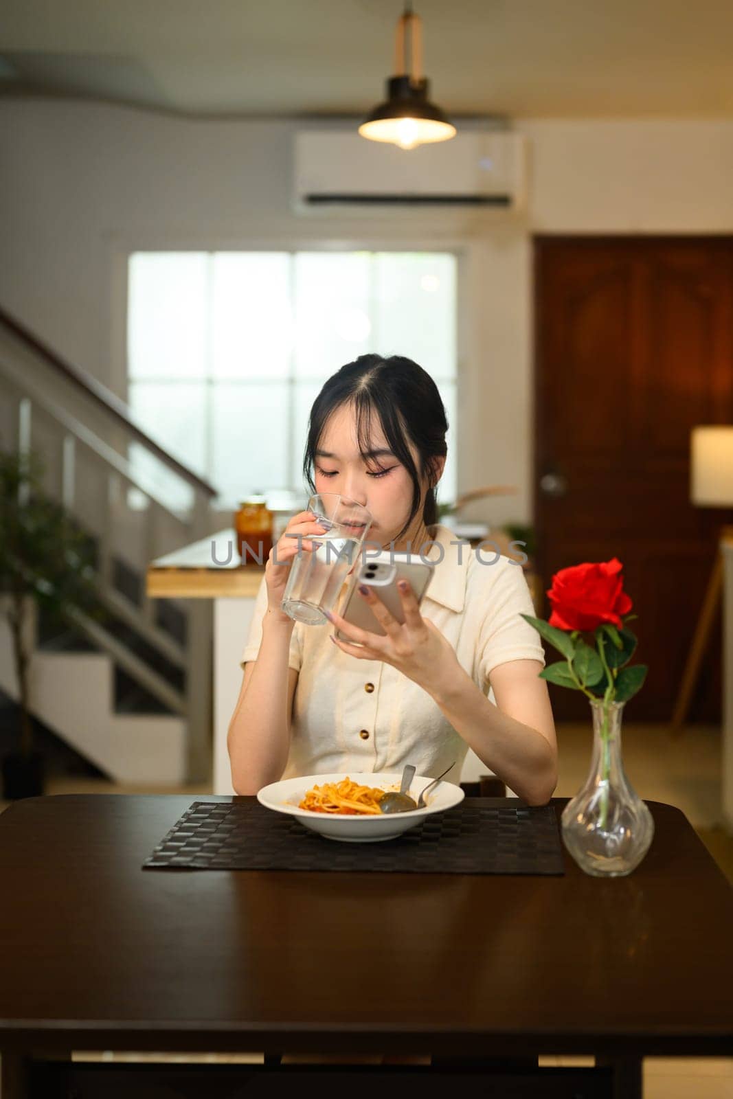 Young woman drinking a glass of water and using her smartphone at the dining table.