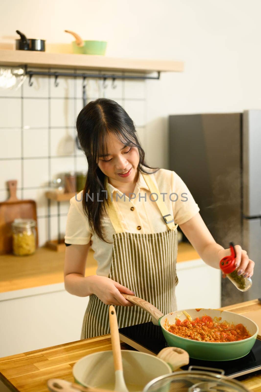 Smiling young woman adding spices into a simmering pan of tomato based spaghetti sauce.