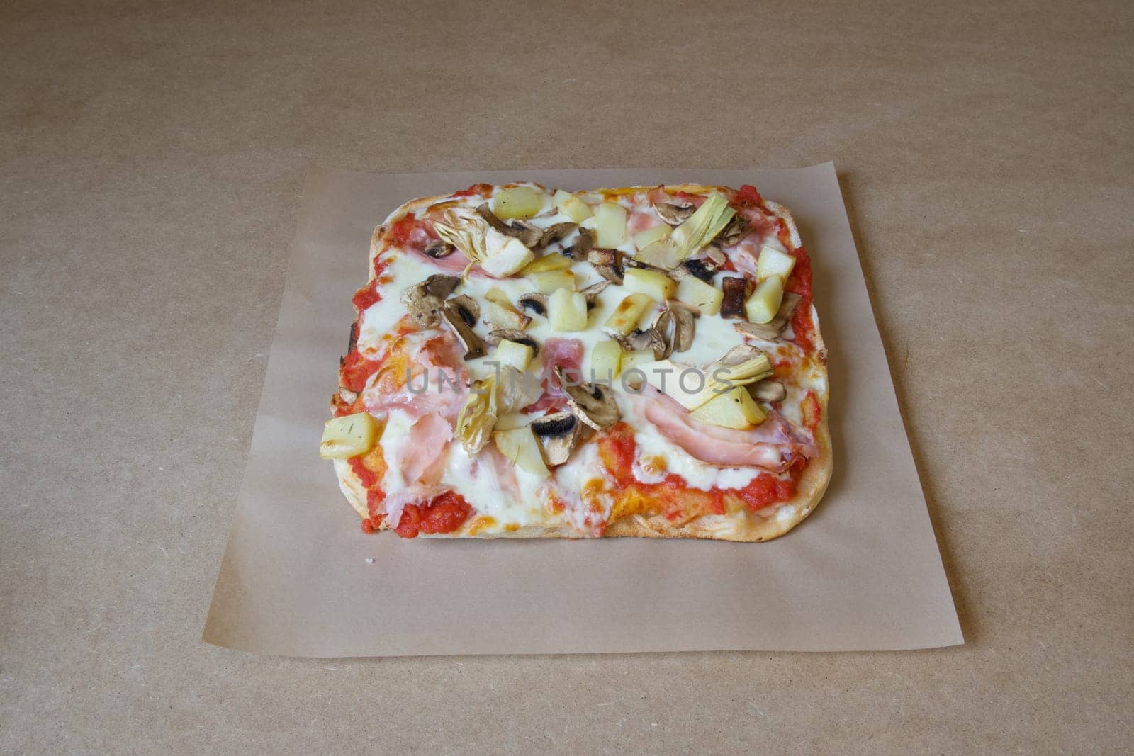 A pizza with ham, mushrooms, and pineapple sits on a brown table. The pizza is cut into a square shape and is covered with cheese