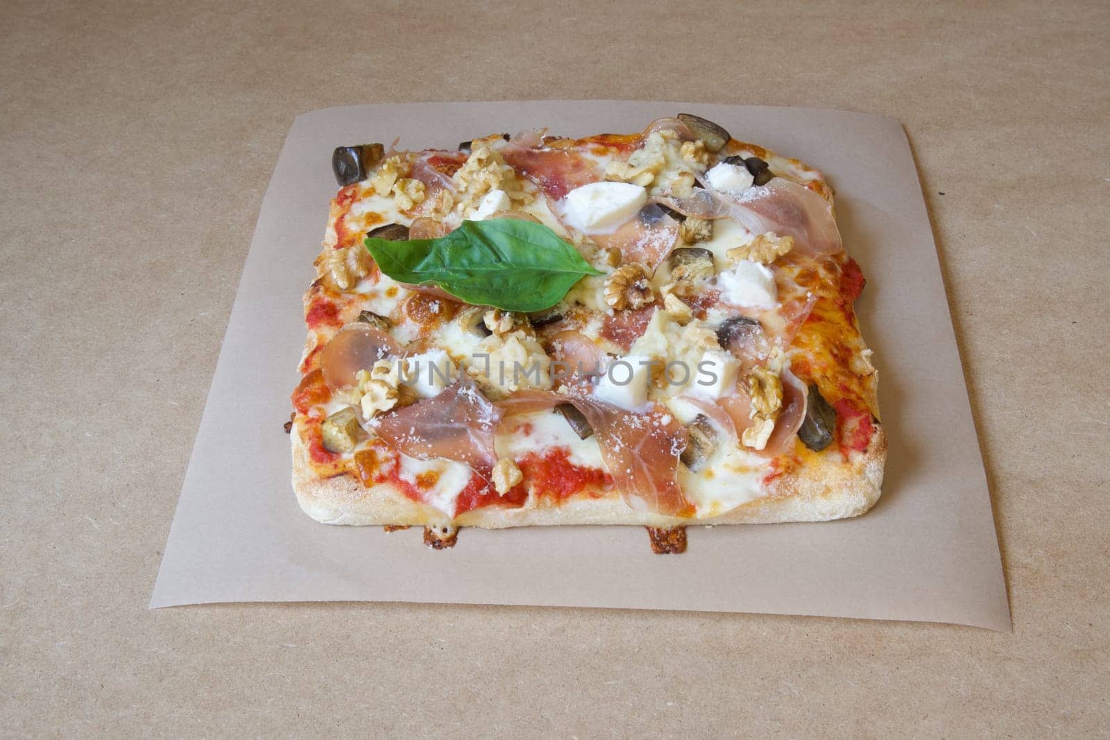 A pizza with walnuts and cheese sits on a brown table. The pizza is cut into squares and is topped with basil