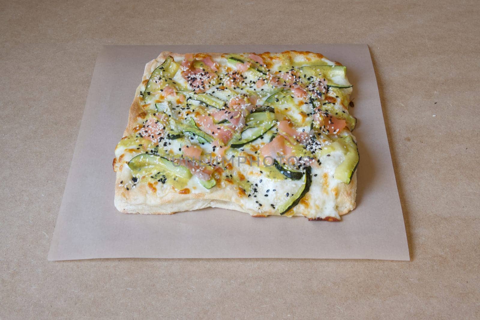 A pizza with vegetables on it is sitting on a brown table. The pizza is cut into squares and has a variety of toppings including cucumbers, tomatoes, and cheese