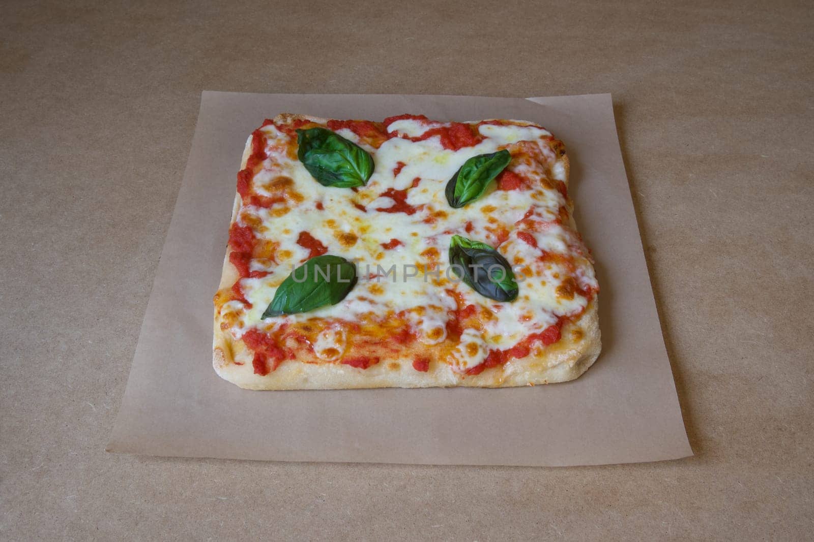 A pizza with basil leaves on top of it. The pizza is sitting on a brown paper. The pizza is cut into squares