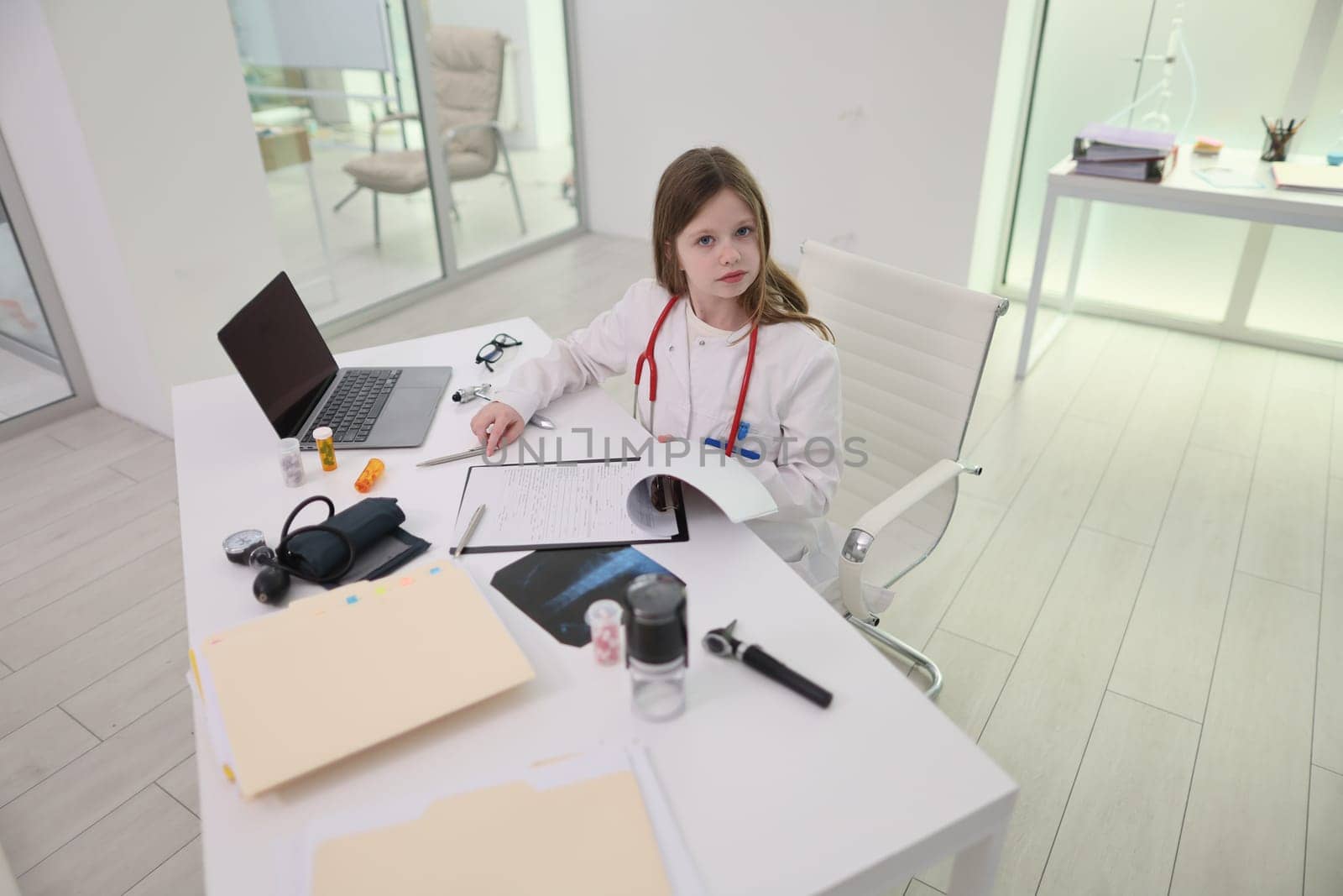 In a chic, minimalist medical office, a young girl dressed as a doctor intently examines a book with great curiosity