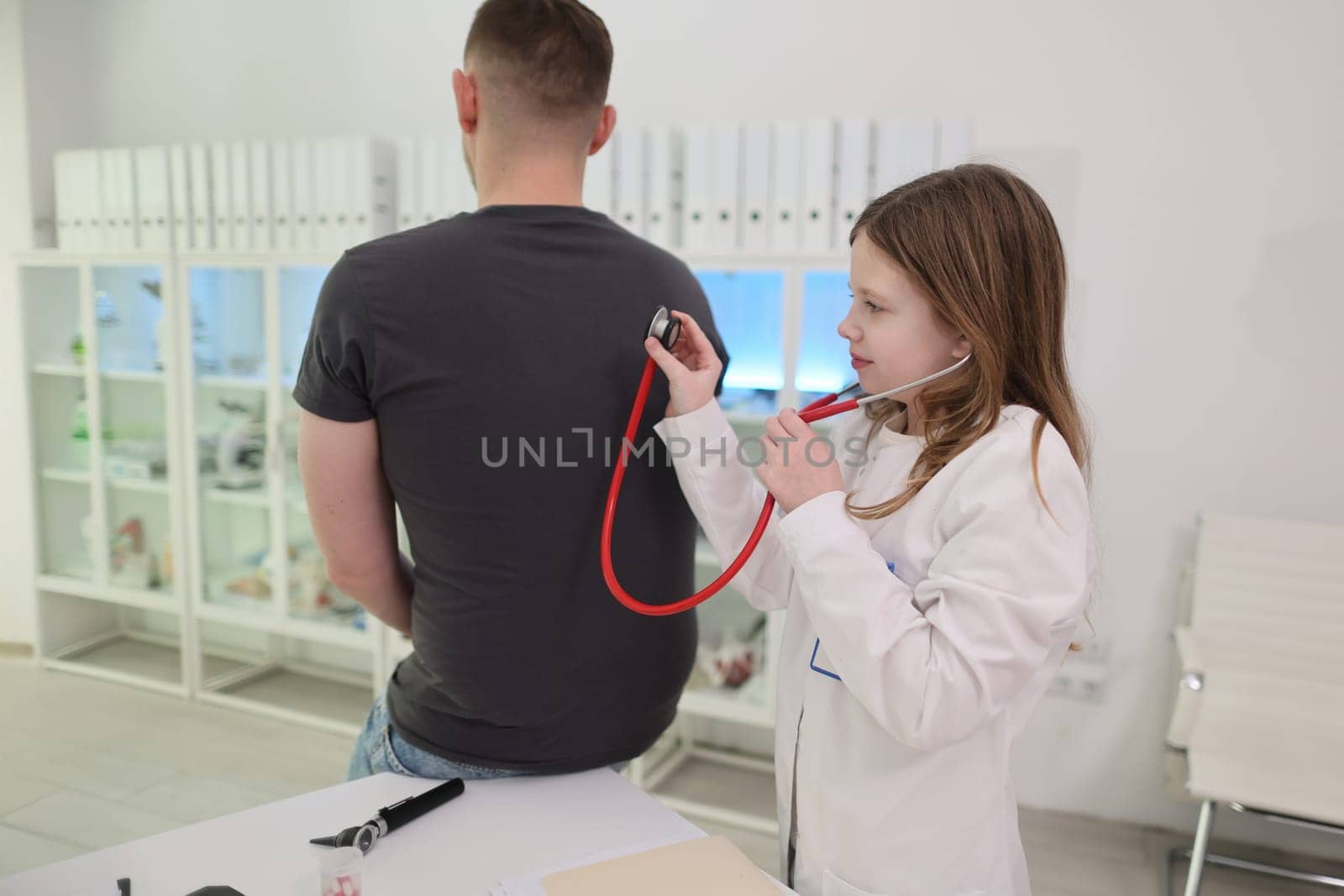A young girl uses a stethoscope to pretend to examine a patient's back in a playful healthcare scenario.