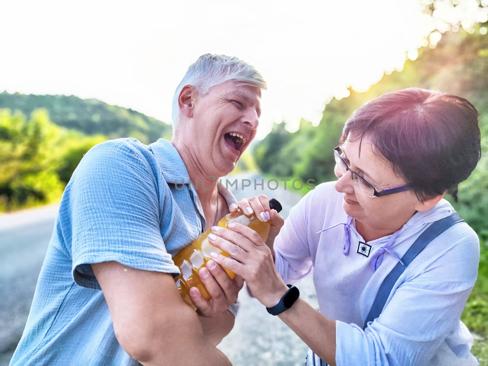 A couple laughs and playfully tugs a drink while walking along a scenic, nature-filled road.