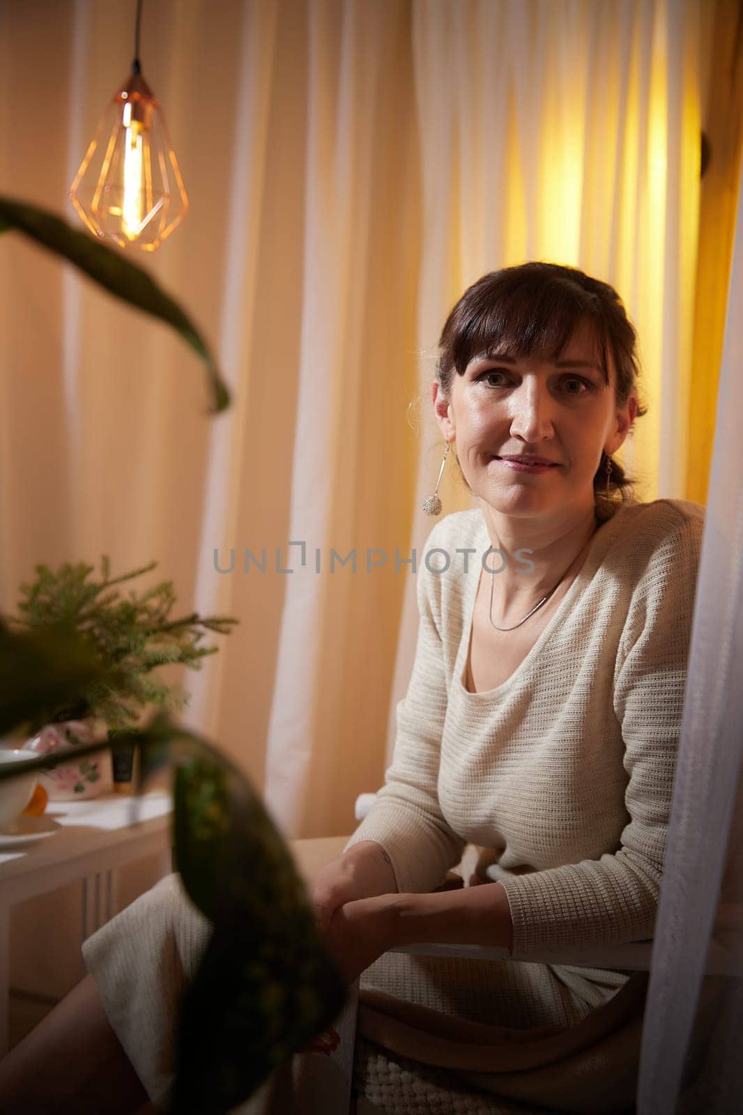 Middle-aged woman sits comfortably in her cozy apartment, enjoying a peaceful evening surrounded by warm lighting and plants.