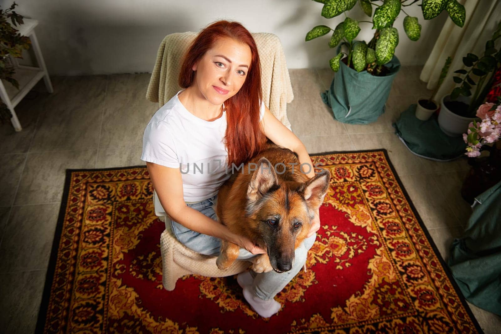 A woman sits comfortably on a chair, gently holding her German Shepherd dog on a patterned rug surrounded by greenery.