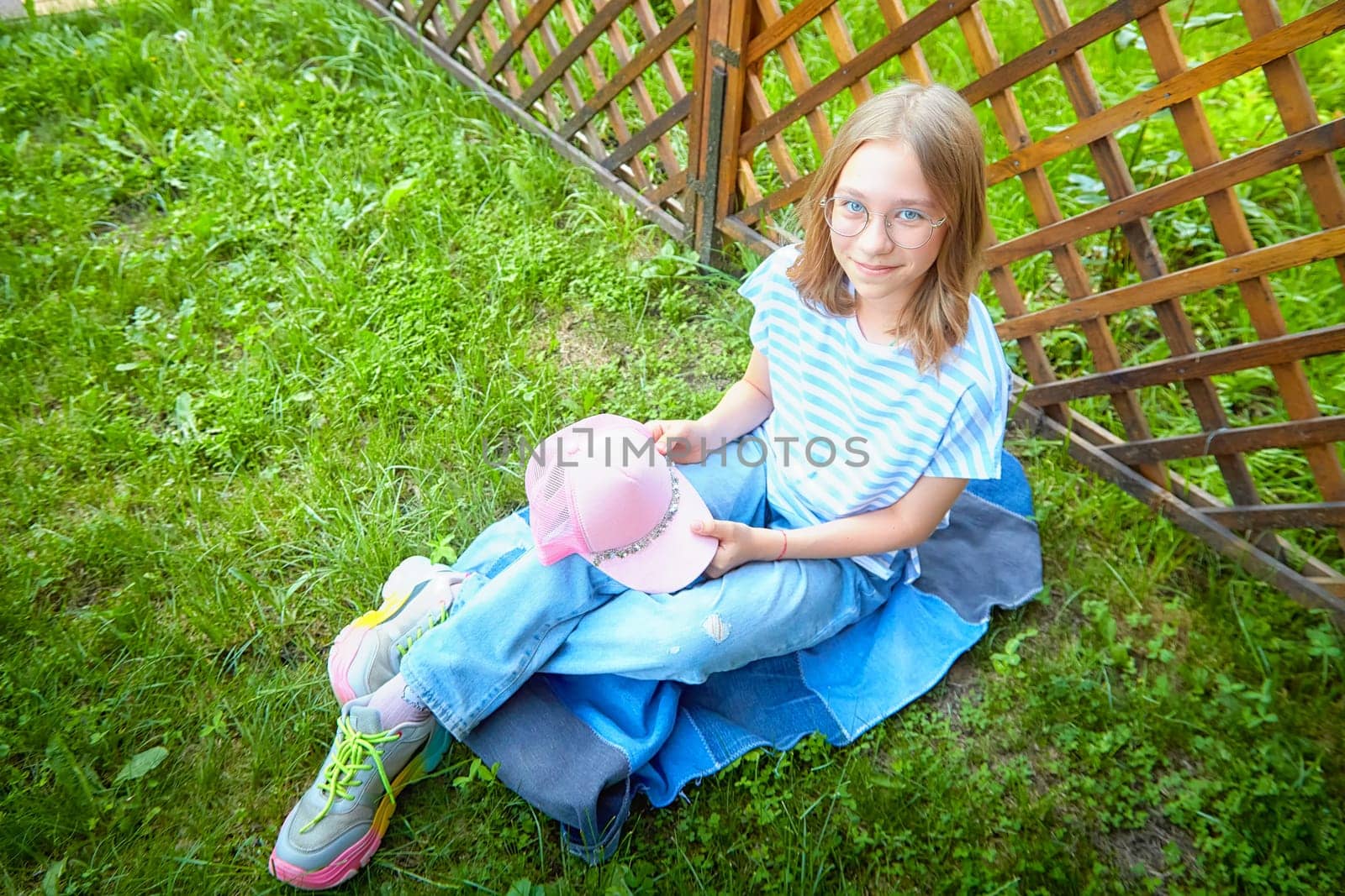Young girl relaxes on grass in comfortable clothing, holding a pink cap, enjoying a sunny day outdoors near a fence.