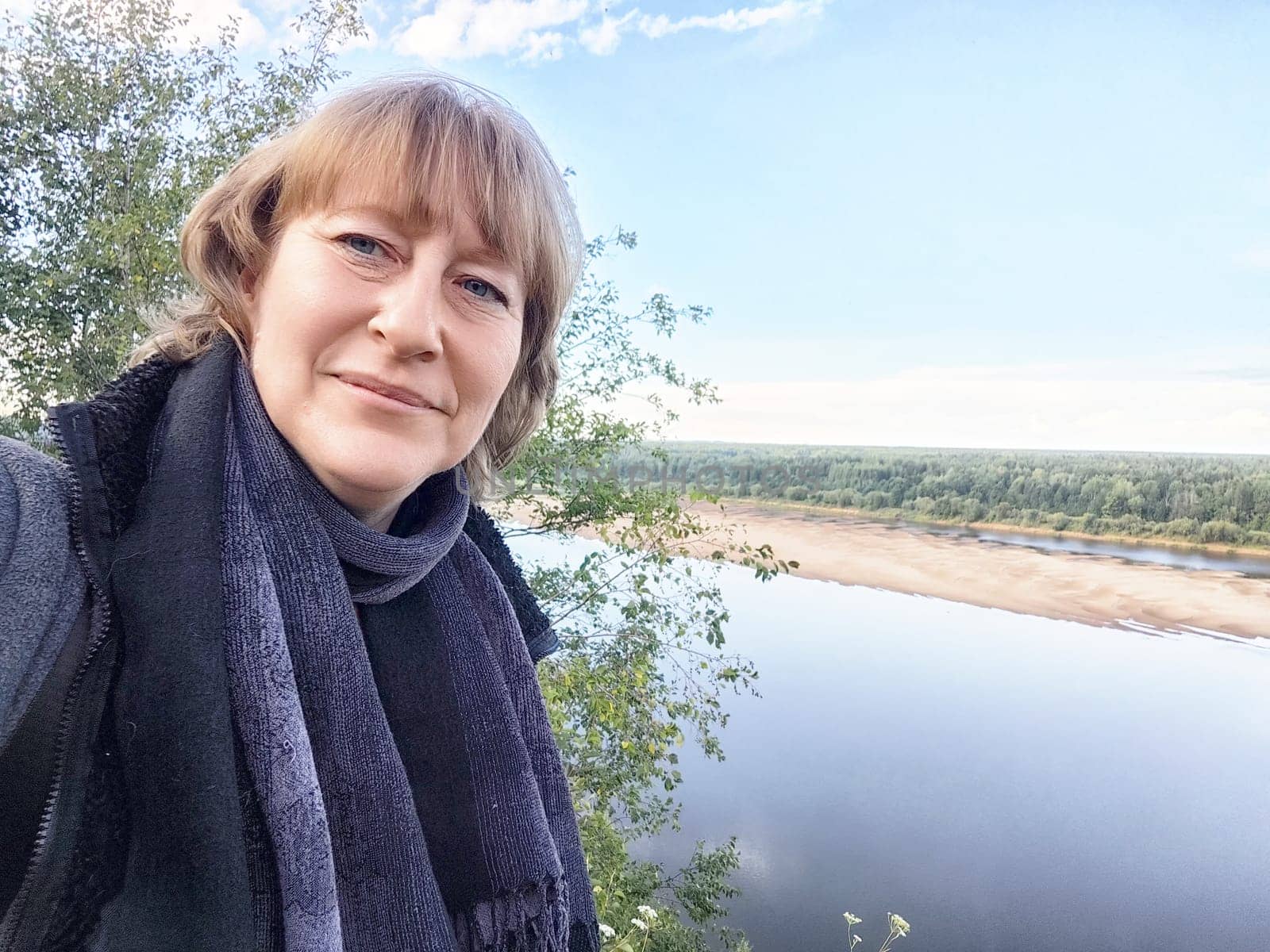 A middle-aged female traveler blogger captures a joyful selfie with a beautiful riverside view in the background.