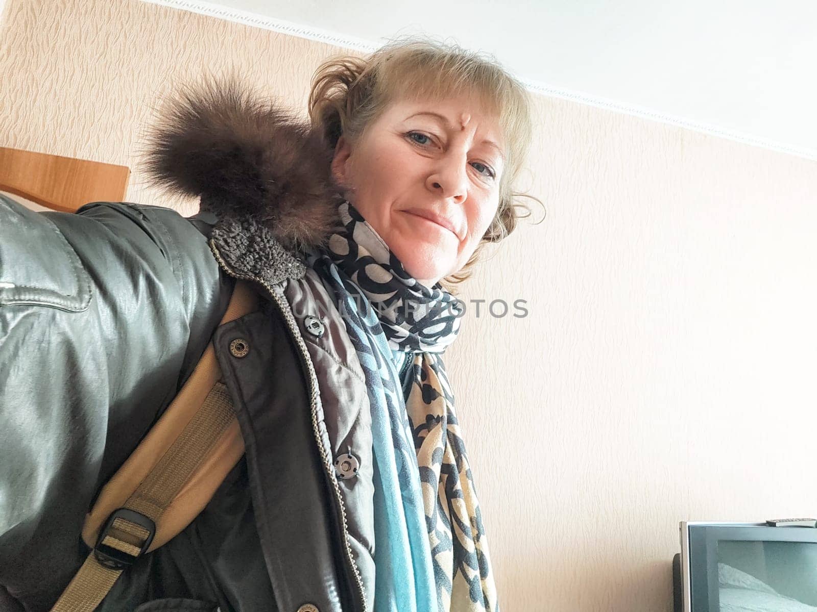 A woman smiles while taking a selfie in a warm winter coat and scarf, standing indoors with a simple backdrop.