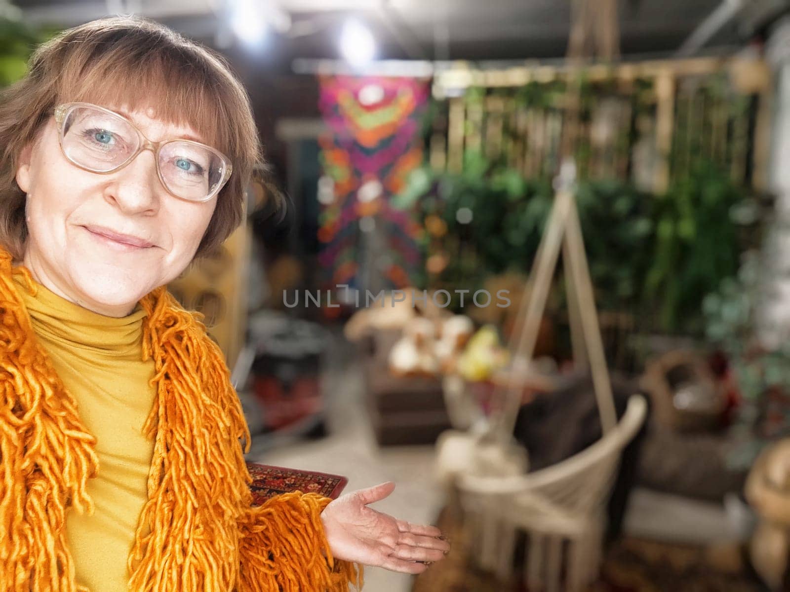A mature blonde woman smiles while gesturing towards her colorful living room, sharing her lifestyle with followers.