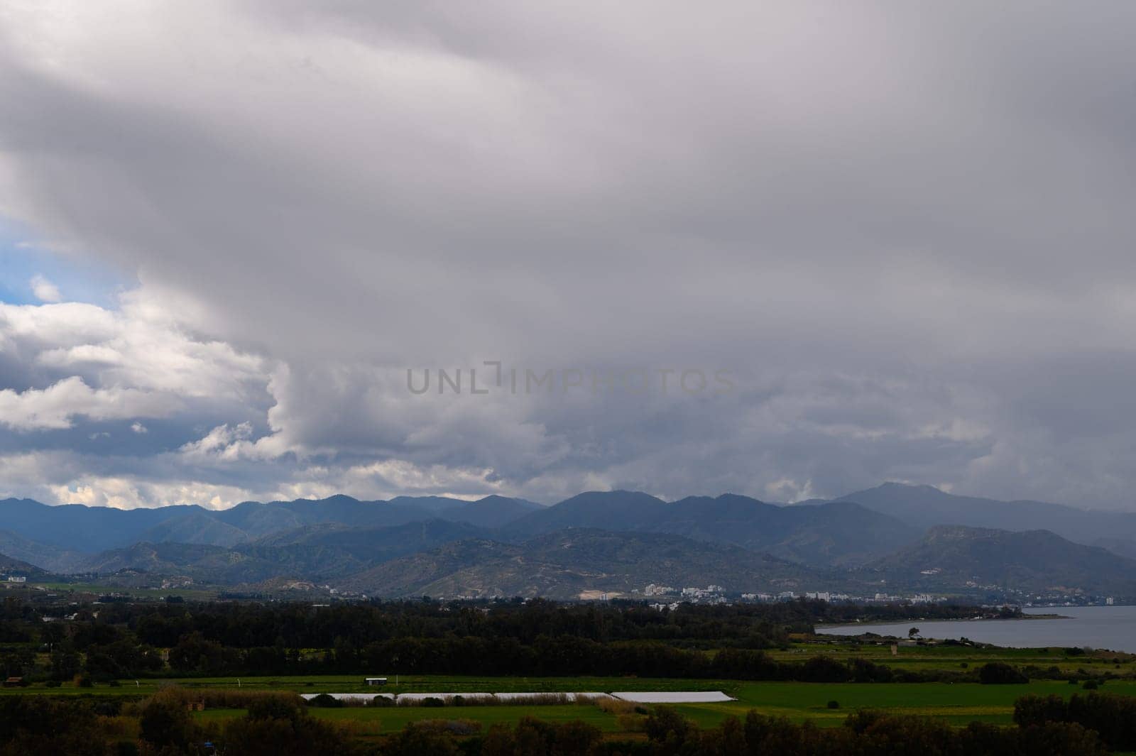 Dramatic clouds billow above vibrant fields while serene water reflects distant mountain silhouettes under gray skies.