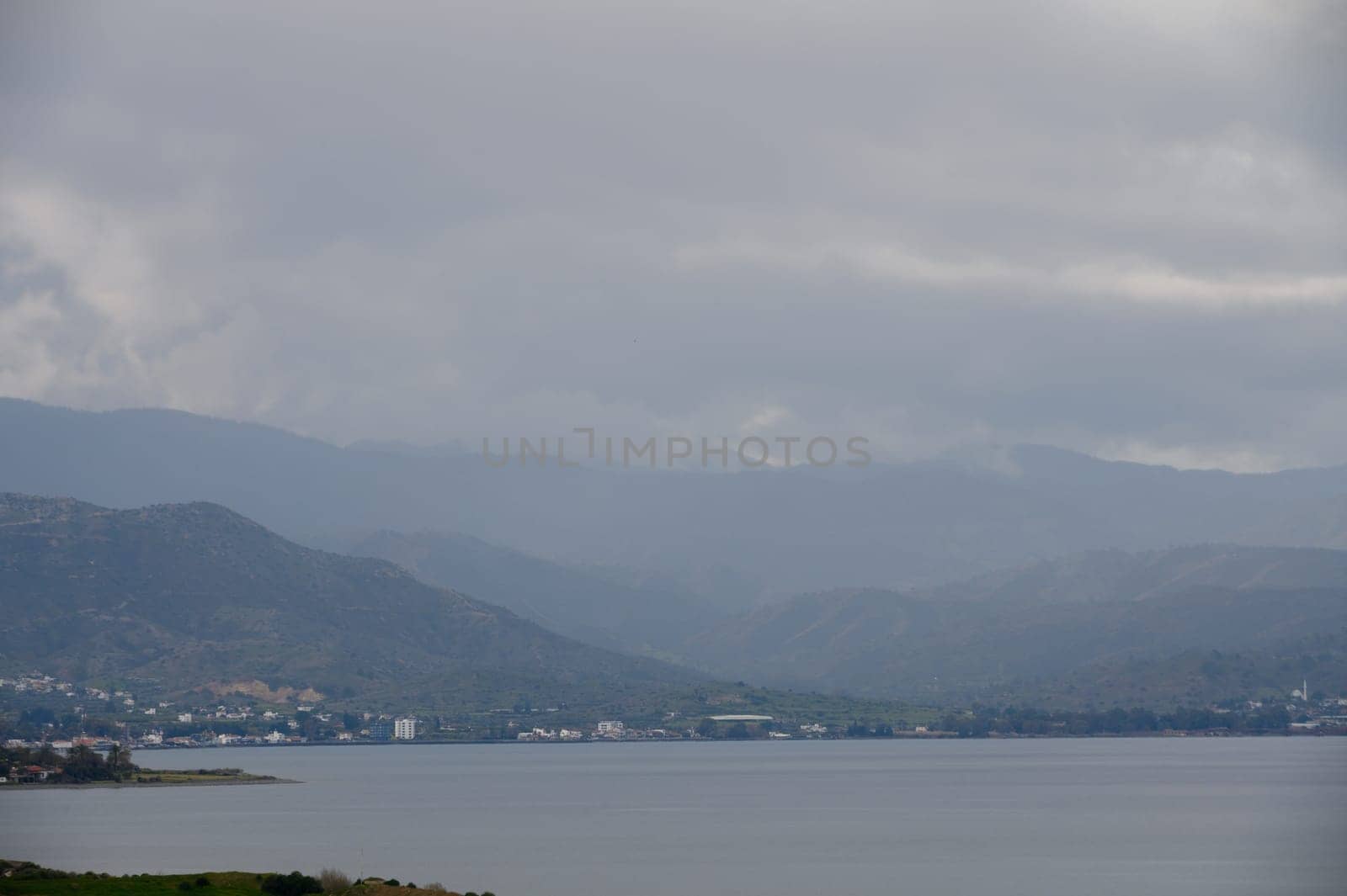 A tranquil lake reflects soft hues as towering mountains linger in the background, shrouded in mist and clouds.