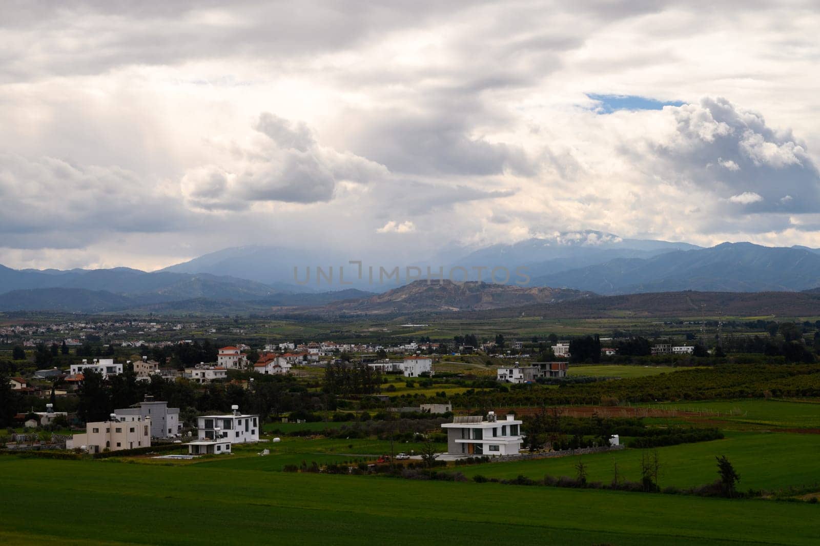 Vast green fields stretch beneath a dramatic sky, framed by distant mountains and modern homes dotting the landscape.