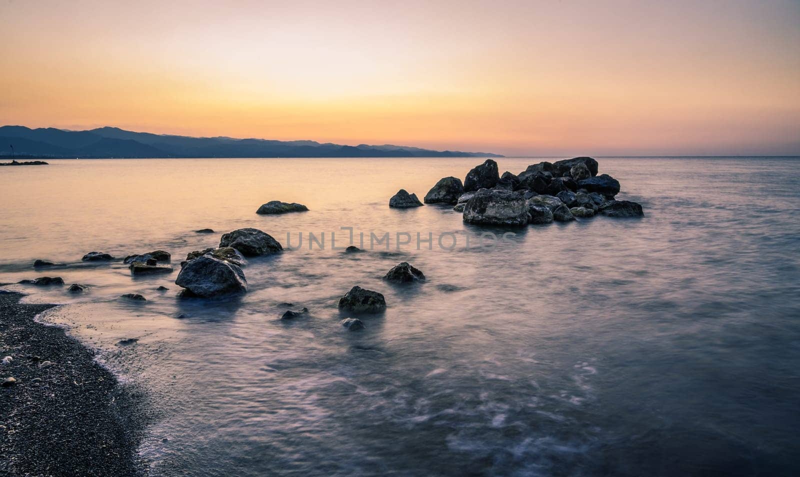 Soft waves lap against rocky shores under a breathtaking sunset, casting warm hues across the horizon.