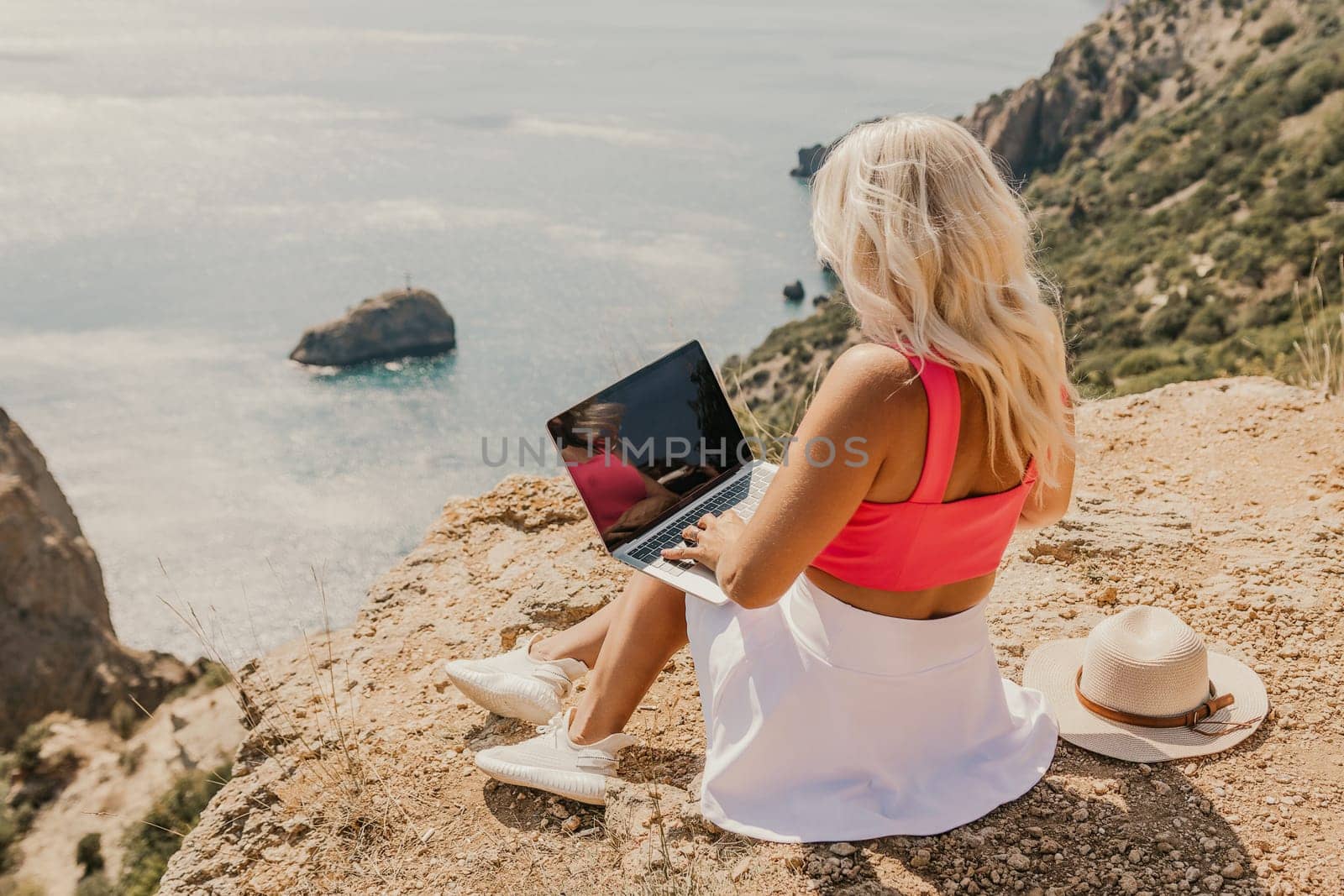A woman is sitting on a rocky hillside with a laptop open in front of her. She is wearing a pink top and a straw hat. Concept of relaxation and leisure
