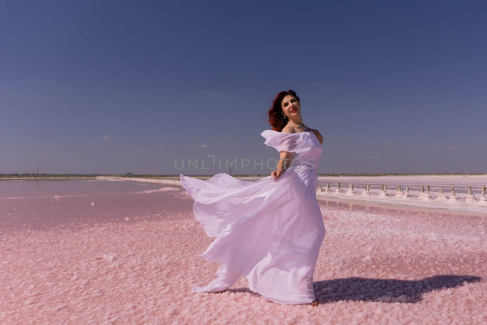 A woman in a white dress walks on a pink salt flat with the wind blowing her dress. The sky is clear blue.