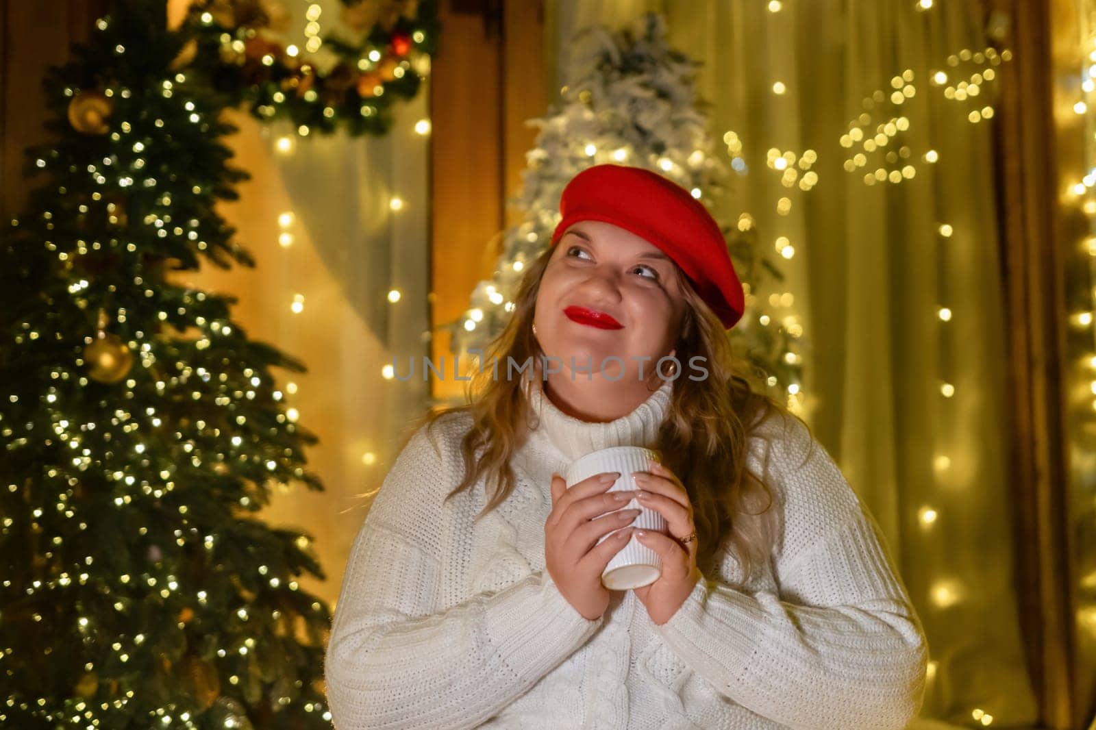 A woman in a white sweater and red beret holds a white mug in front of a Christmas tree with string lights.
