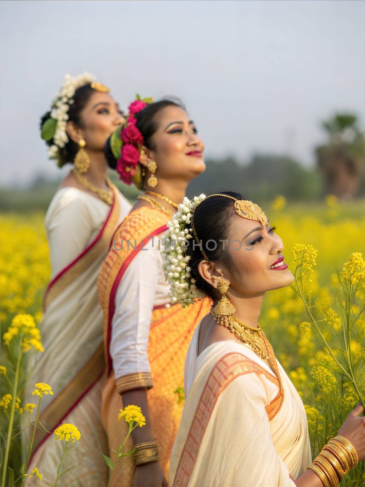 Assamese women in elegant Mekhela Chador, adorned with traditional gold jewelry and fresh flowers in their hair, standing in a yellow mustard field