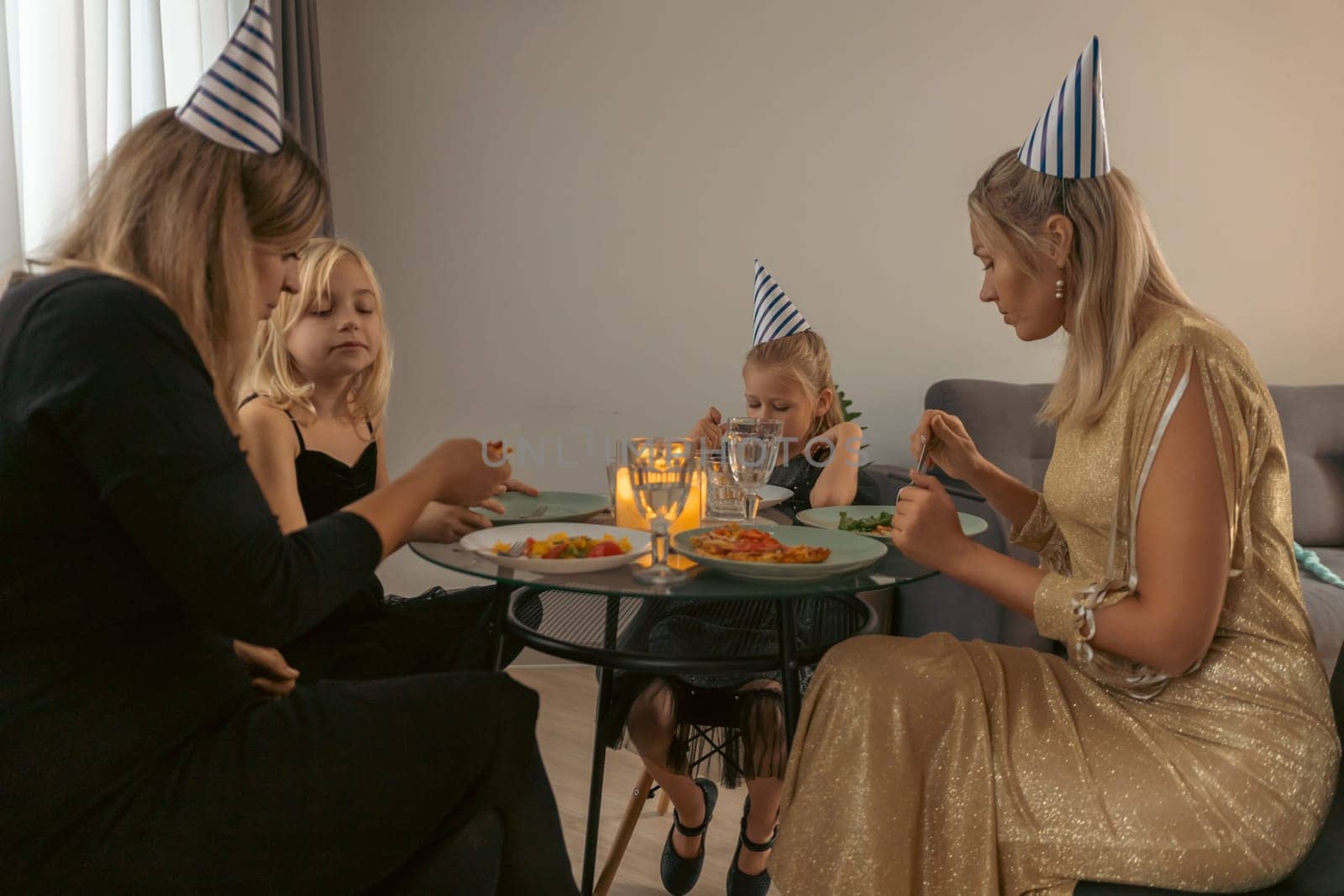 Two women and two girls, wearing party hats, sit around a table, enjoying a meal with lit candles. A small birthday celebration indoors.