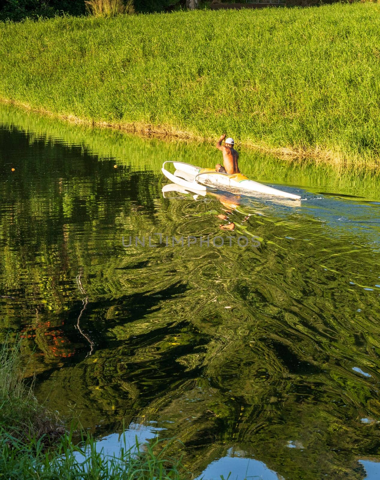 A man paddles a traditional Hawaiian outrigger canoe through the tranquil waters of Hanalei River on a sunny day, creating mirrored reflections.