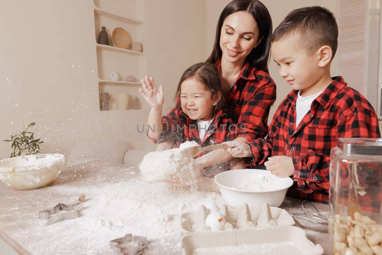 Happy mother and kids baking together, playing with flour in a cozy kitchen, wearing matching plaid shirts, joyful family bonding and messy fun moment.