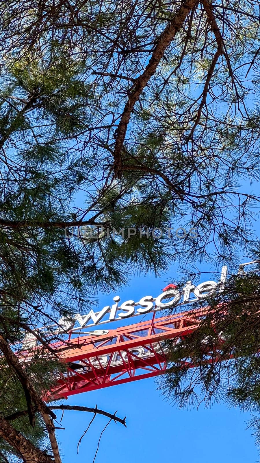 22.02.2025.Budva,Montenegro.Swissotel sign on crane boom partially visible behind tree branches, with red structure against clear blue sky.