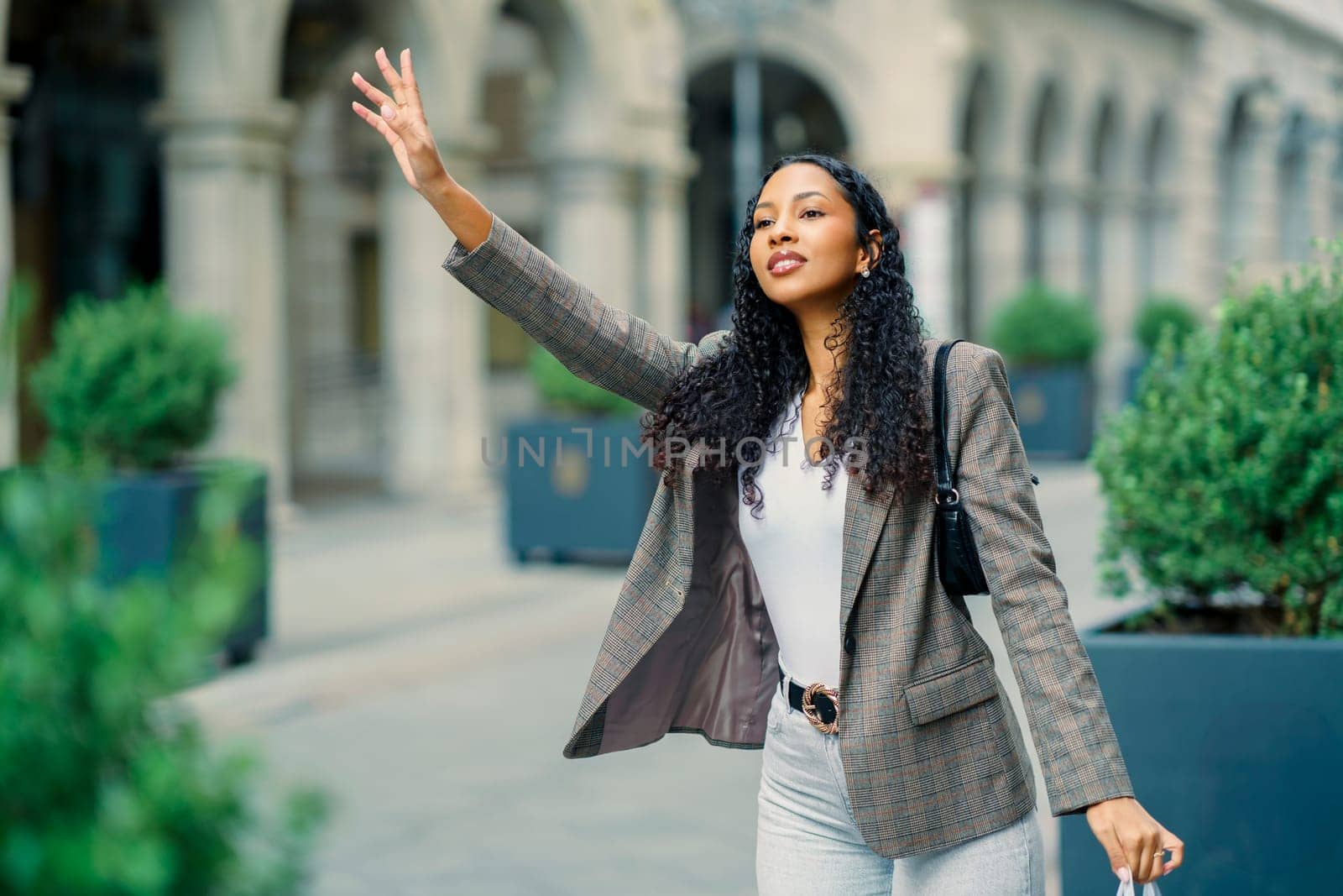 A stylish young woman with beautiful curly hair waves cheerfully while walking confidently through a vibrant urban area, showcasing her impeccable fashion sense and lively personality with every step