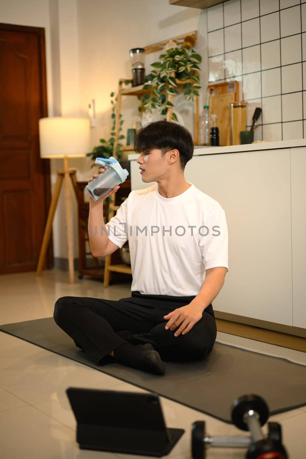 Handsome man sitting on yoga mat, drinking a protein shake after a workout.