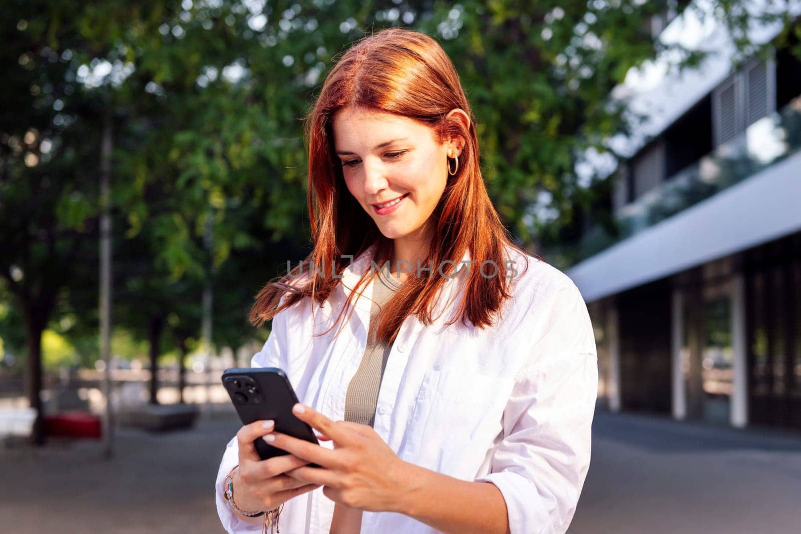 smiling woman using cell phone while walking down the street, she is wearing a white shirt and is focused on his phone, concept of technology of communication and urban lifestyle, copyspace for text