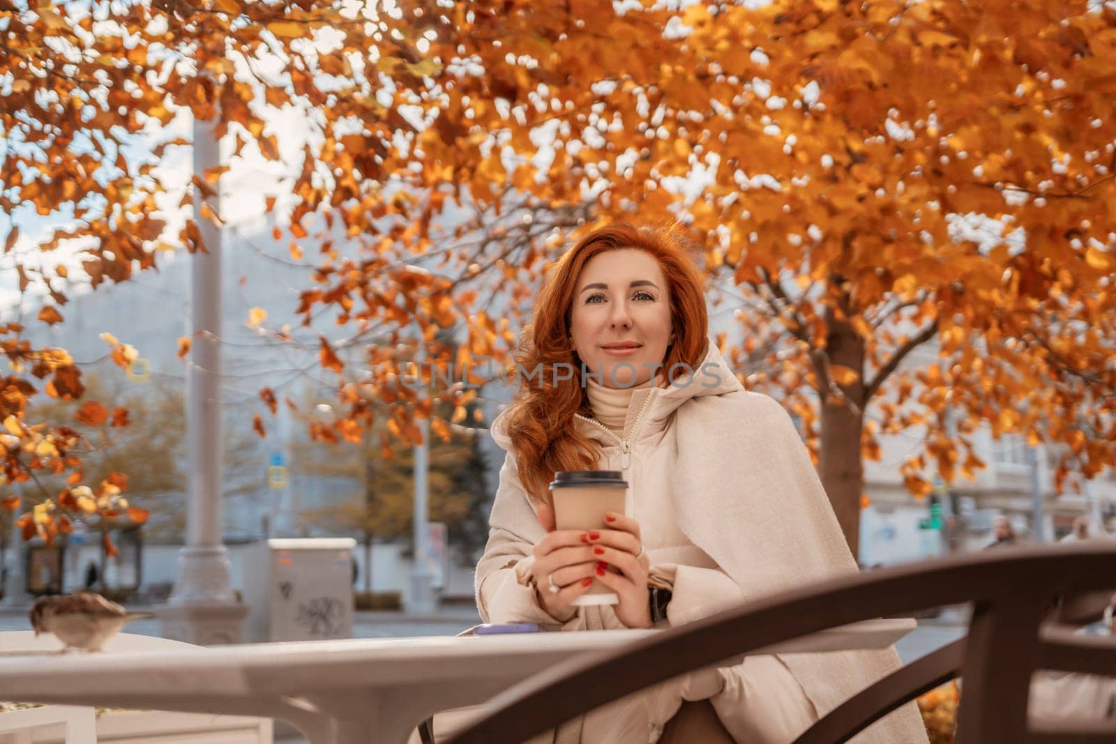 Redhead woman smiles, holding a coffee cup, seated outdoors under vibrant autumn leaves on a park bench.