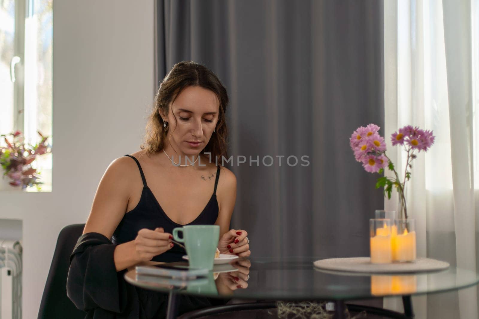 Woman sits at glass table, sipping from teal mug. Dark clothing, flowers and candles nearby. She appears thoughtful.